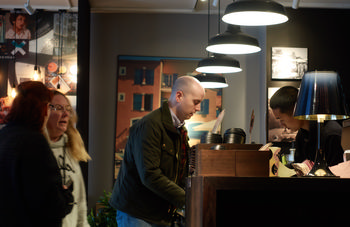 Coffee shop order This indoor urban photograph captures a coffee shop scene in Copenhagen during a morning in autumn. The main subject of the image is a coffee shop order being placed at the counter by people dressed in seasonal clothing, suggesting cooler weather. Several customers are interacting in front of the wooden counter, which is equipped with stacked takeaway cups and utensils. Industrial-style hanging lights illuminate the interior, creating a warm atmosphere, while framed images and a large photograph of Copenhagen architecture decorate the walls. The composition highlights everyday life in the city, with people engaged in typical morning routines. The photograph is by Jamie Weinbren.